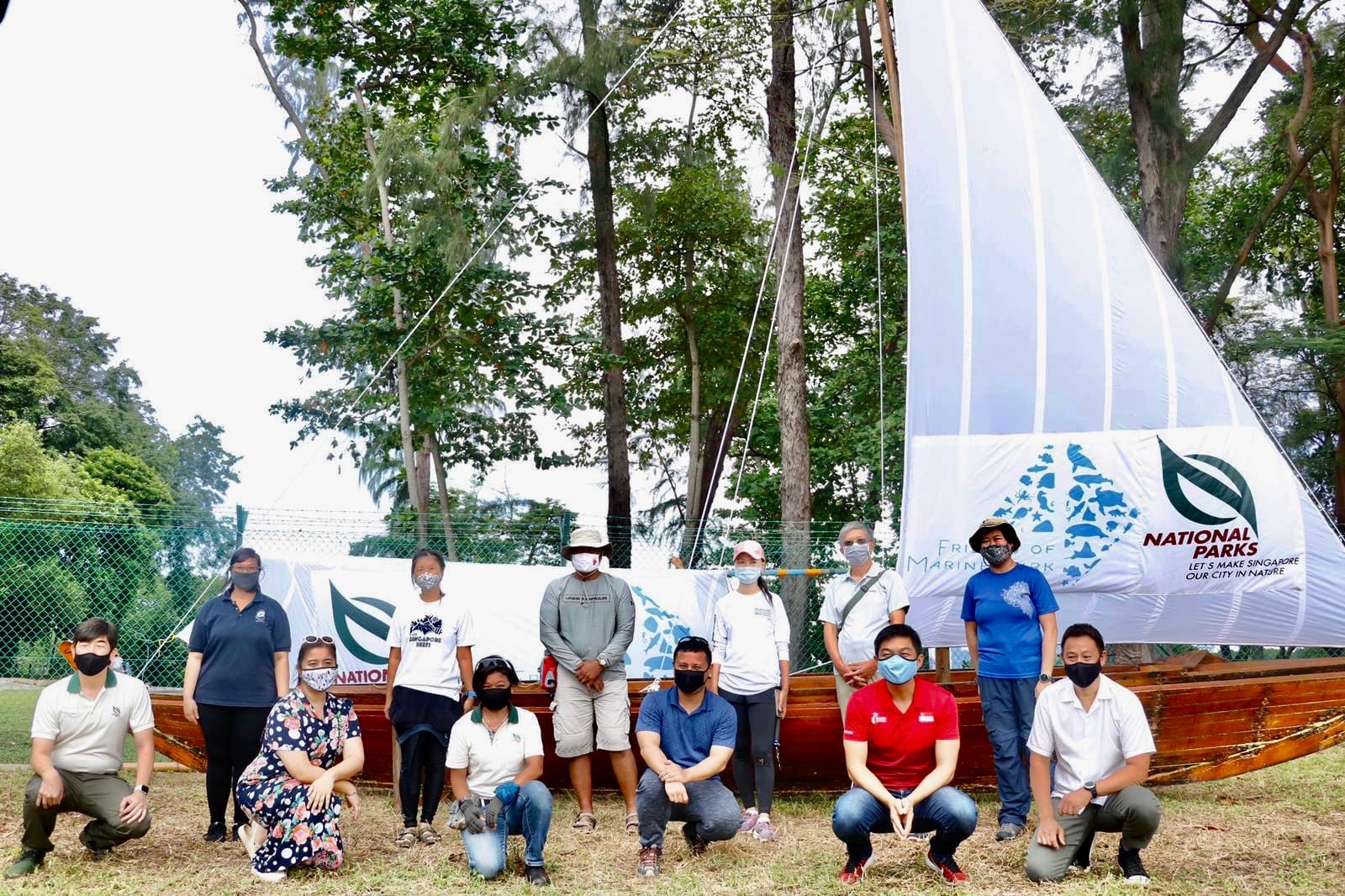 Group of people standing and kneeling in front of a wooden sailboat with a "National Parks" banner in a wooded area.
